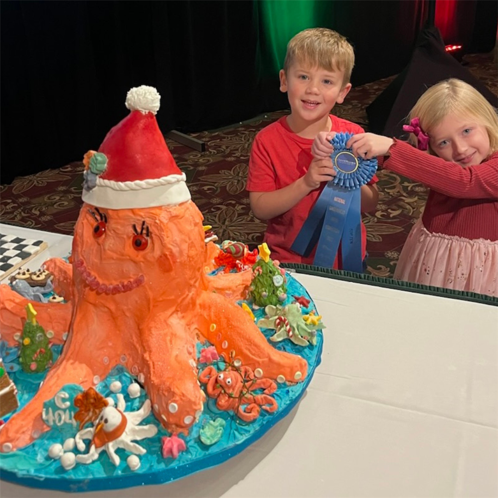 boy and girl hold award ribbon behind orange gingerbread octopus with Santa hat.